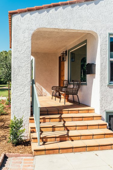 A Spanish white house with black wire chairs on the porch, terracotta steps, and terra-cotta tile roof