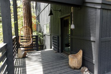A wood deck of a black cabin. A hanging boho chair, wood stools, and black lamp sconce.