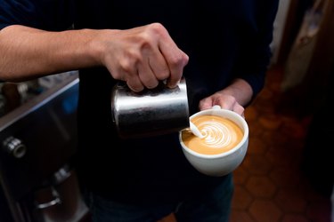 A person pouring hot milk into a cup of coffee, forming a swirl pattern for a latte.