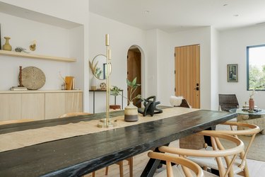 Black dining table with beige table runner, candlesticks, twisted globe sculpture. A light wood sideboard with sculptures and a wood door.