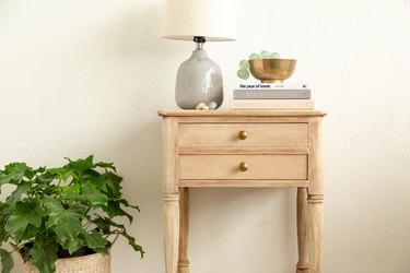 Wood side table with double drawers, gold knobs, spindle legs, with a lamp, books, bowl of beads, and a plant below