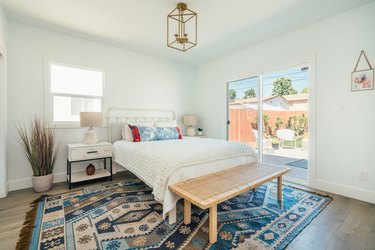 An ornate blue-beige rug, white bedding with colorful pillows, two houseplants, large picture window, and gold ceiling lantern