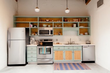 Kitchen with light blue-wood cabinets, art deco pendant lights, and an ornate wall grille vent, and purple-pink floor rug.