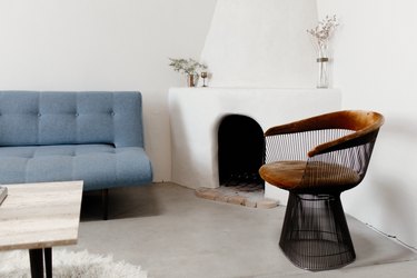 Minimalist living room with a brown-black wire modernist chair. White Adobe fireplace with vases of dried flowers. A blue tufted sofa.