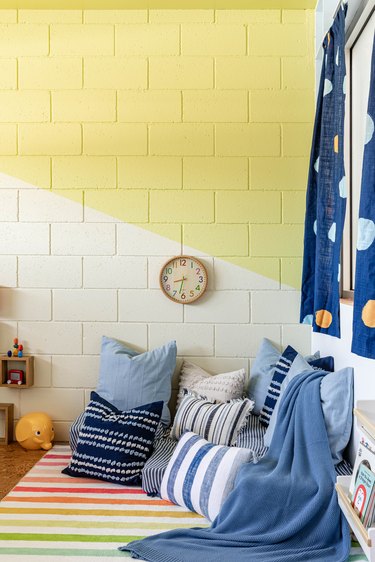Kid's bedroom corner with yellow walls, blue throw pillows, rainbow striped rug, and wood frame wall clock