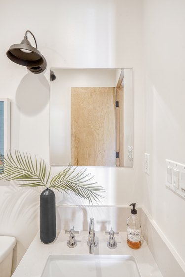Bathroom sink with a black oblong vase of palm leaves. A rectangular mirror with a black bell sconce light.