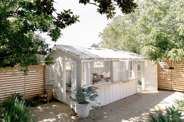 White greenhouse with a corrugated roof and open windows with wood counters. Wood fence and trees surround.