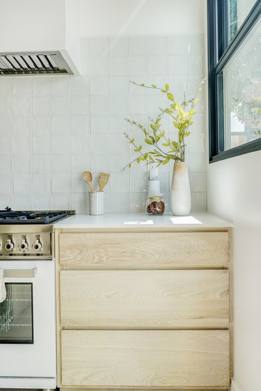 A light wood kitchen cabinet with vases of flower clippings and a vase of wooden utensils all beside a stovetop