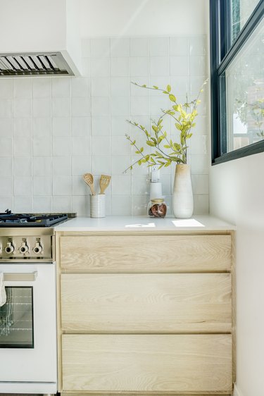 A light-wood kitchen cabinet with vases of flower clippings and a vase of wooden utensils all beside a stove top