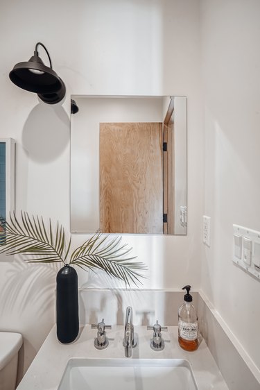 Bathroom with a porcelain sink, metal faucets, black vase with palm leaves, black bell farm sconce light, and a rectangular mirror