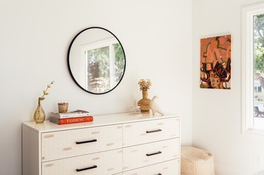 A white dresser with a beige vase and bird sculpture with a round mirrorr and contemporary wall art