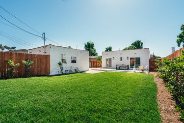 A grass lawn with plants bordering a wood fence enclosing a Spanish style house with terra-cotta roofing