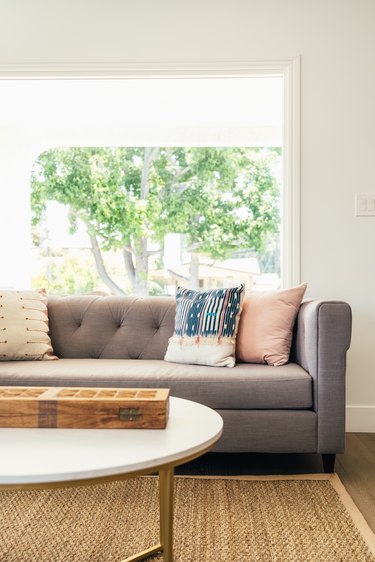 Gray living room sofa with multi-colored pillows by a picture window. A white round coffee table on a neutral fiber rug.