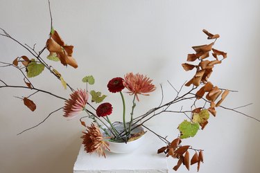 Floral ikebana arrangement with pink and red flowers, and branches of leaves, all on a plinth