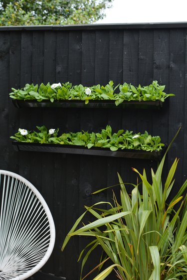 Black DIY gutter garden attached to black-painted wood fencing with white patio chair and grassy plant