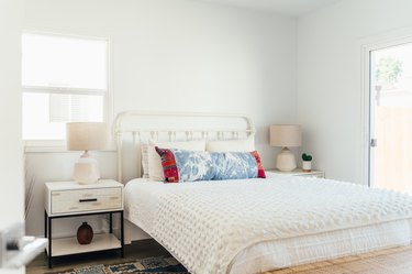 A bedroom with white bedding and blue-red pillows, symmetrical white nightstands and lamp