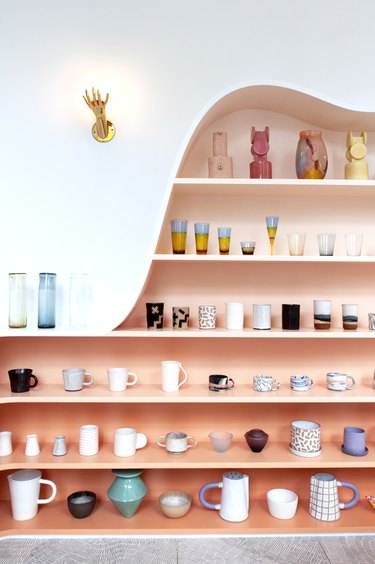 Pink display shelves with a wavy top and eclectic dish ware, and a hand shaped sconce.