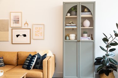 Gray cabinet with an arched top, inside are white vases, plants, and books and a beige sofa with blue pillows.