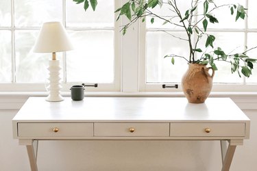 Home office corner with a white desk by a window, accented with a lamp, vase of leaf cuttings, and a mug