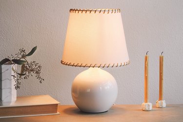SKOTTORP lampshade with leather whipstitching, on a table with candlesticks, a book, and a vase of flowers