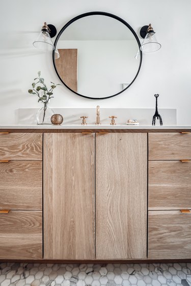 Wood vanity drawers, gray tile floor, round mirror, wall sconces, plant clipping, and small sculpture in a bathroom