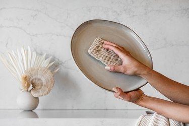Person cleaning an oval dish with an eco-sponge. A vase of dried florals is on a marble or granite countertop.
