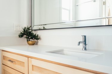 Light wood bathroom vanity with a white countertop, metal sink faucet, and a gold bowl with a plan clipping all underneath a mirror