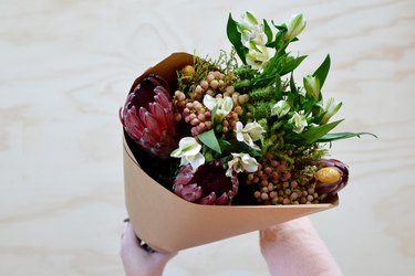 Hands holding bouquet with white, pink, red and yellow flowers with green leaves wrapped in brown paper against a light wood background