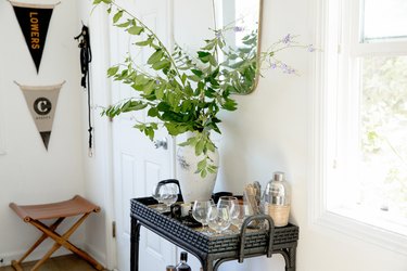 Black bar cart with glasses, cocktail shaker, and vase of leaf cuttings. Brown canvas stool and flag decor