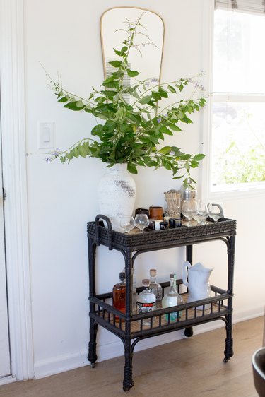 Black bar cart with bottles, glasses, and plant clippings in a vase. Window and an oblong mirror.