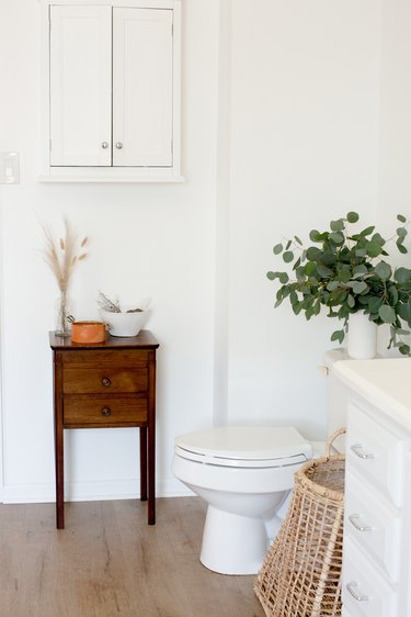 white toilet, small side table with drawers, rattan basket, vase filled with eucalyptus, white wall-hung cabinet, wood flooring, white bathroom vanity