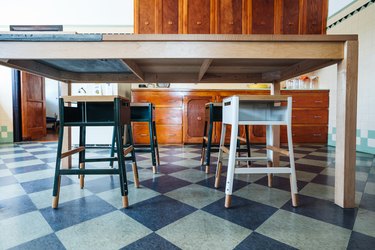 A blue-beige chessboard floor. Square stools under a wood table. Dark wood kitchen cabinets. Green-beige tiled walls.