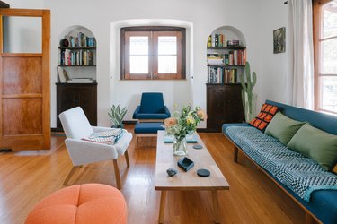 Living room with wall niches, wood sideboards and bookshelves. Wood coffee table with flowers. Blue and orange furniture and pillows.