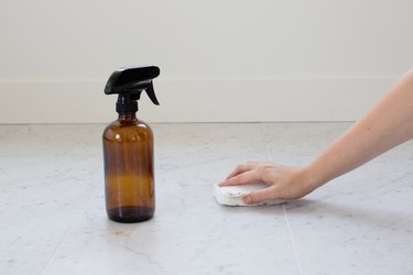 A hand scrubbing a granite or marble tile floor with a sponge. A brown spray bottle is beside.