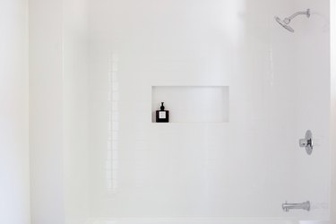 A white subway tile shower. A shower niche is in the wall with a black dispenser bottle. A silver rain shower head and handles are mounted.