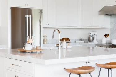 White kitchen island with a marble countertop. Two bar stools with wooden seats and metal legs in fron of it. On the countertop, two white mugs and a chemex, along with a sink with a chrome faucet. Behind the island, a stainless steel fridge and white cabinets.
