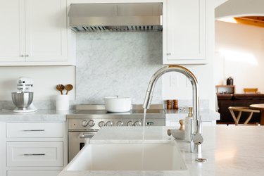 In a kitchen, a sink in a marble countertop, part of a kitchen island. The chrome faucet is running water. In the background, there's a chrome range with a white dutch oven on top.