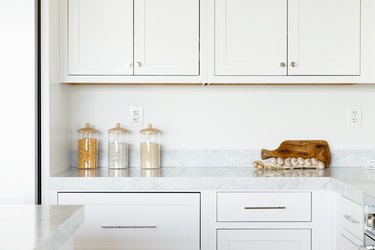 Kitchen counter. The countertop itself is marble, and the cabinets above and below are white. On the counter, three glass food storage containers, a cutting board, and a string of garlic.