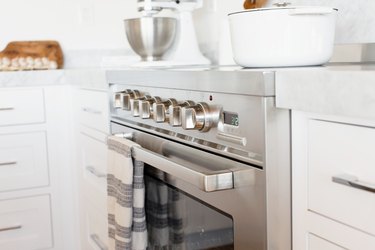 An oven door and temperature control knobs on a stainless steel range. A white and grey striped kitchen towel is hanging over the handle over the oven door.