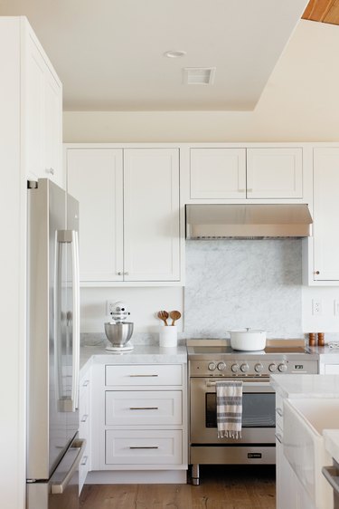 Kitchen with a stainless steel range, white cabinets, stove vent, and a marble countertop. On the countertop, there's a white KitchenAid stand mixer with a stainless steel mixing bowl along with a white utensil holder with wooden salad tongs. On the stovetop, there's a white dutch oven. To the left, there's a stainless steel refrigerator. To the right, there's a white kitchen island with a sink.