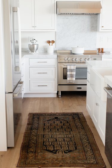 Kitchen with a stainless steel range, white cabinets, and a marble countertop. On the countertop, there's a white KitchenAid stand mixer with a stainless steel mixing bowl along with a white utensil holder with wooden salad tongs. On the stovetop, there's a white dutch oven. On the wood floor, there's a small rug.