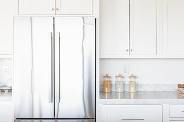 Double doors of a stainless steel refrigerator with white cabinets overhead. Next to it, marble countertop with glass food storage containers and white cabinets.