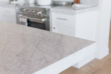 Closeup of corner of marble countertop. In the background, stainless steel range and white cabinets and drawers.