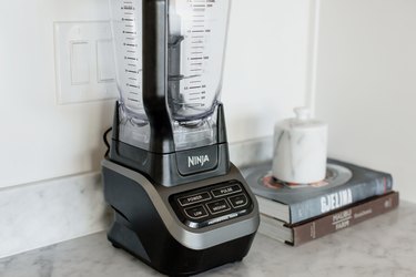 Black Ninja blender on a marble countertop. Next to the blender, two cookbooks – Gjelina and Malibu Farm – are stacked on top of each other. A marble container with a lid is on top of them.