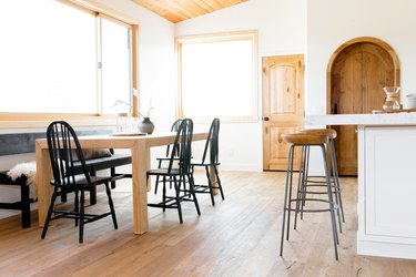 An open interior space, a large dining table on one side and a white kitchen island on the other side. At the table, there are four black Windsor chairs and a black wood bench, which is against the wall. Three wooden bar stools with metal legs sit in front of the kitchen island. There are two windows lining the white walls and two wooden doors.