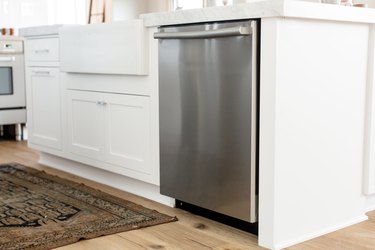 Stainless steel dishwasher in a white kitchen island. The marble countertop extends over the dishwasher. On the floor, a small rug.