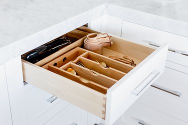 An open drawer with a wooden cutlery organizer. Cutlery, measuring cups and spoons, and other utensils visible inside. Marble countertop above the drawer.