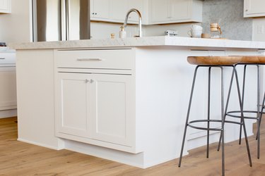 A row of bar stools with wooden seats and black metal legs in front of a white kitchen island with a marble countertop. Visible on the countertop is a chrome faucet, two stacked white mugs, and a Chemex.