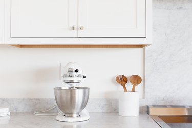 White cabinets above a marble counter. On the counter there is a white KitchenAid stand mixer with a stainless steel mixing bowl. Next to the mixer is a white utensil holder with wooden salad tongs.