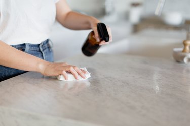 Shot of the midsection and hands of a woman in a white t-shirt and jeans. She's wiping down a kitchen counter with a brown glass spray bottle in one hand and a white rag in the other.